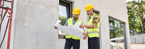 Devoted builders in safety vests and helmets looking at blueprint while working outside, banner — Stock Photo
