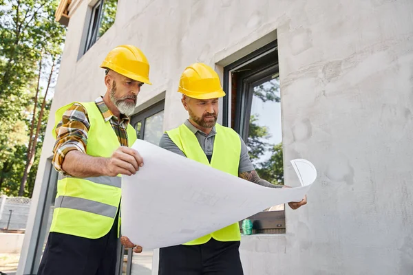 Good looking bearded builders in safety vests and helmets looking at blueprint while working outside — Stock Photo