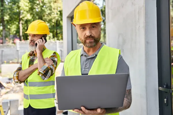 Construtor atraente com tatuagens trabalhando no laptop enquanto seu colega falando por telefone durante o trabalho — Fotografia de Stock