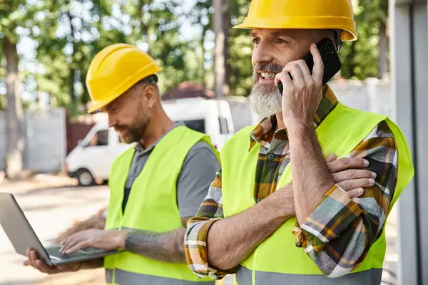 Attractive builder with tattoos working on laptop while his colleague talking by phone during work — Stock Photo