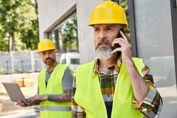 Attraente costruttore con tatuaggi che lavorano sul computer portatile mentre il suo collega parla per telefono durante il lavoro — Foto stock