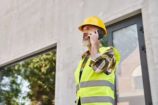 Joyeux homme dévoué dans le casque de sécurité parler par téléphone sur le chantier de construction, constructeur de chalet — Photo de stock