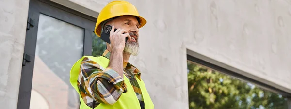 Alegre hombre dedicado en casco de seguridad hablando por teléfono en el sitio de construcción, constructor de cabañas, pancarta - foto de stock