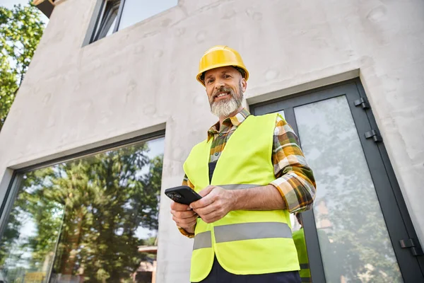 Bell'uomo gioioso in giubbotto di sicurezza e casco in posa con il telefono e guardando la fotocamera, costruttore — Foto stock