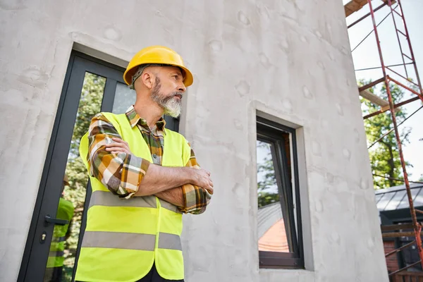Hombre barbudo guapo en chaleco de seguridad y casco mirando hacia otro lado mientras que en el sitio de construcción, constructor - foto de stock