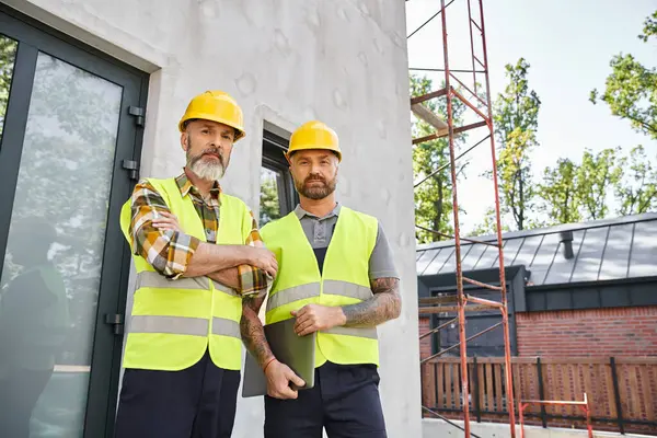 Good looking devoted men in safety attires with laptop looking at camera while outdoors, builders — Stock Photo