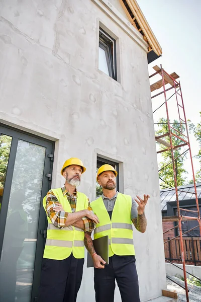 Dos trabajadores de la construcción de buen aspecto en chalecos de seguridad y cascos discutiendo sitio, constructores de cabañas - foto de stock