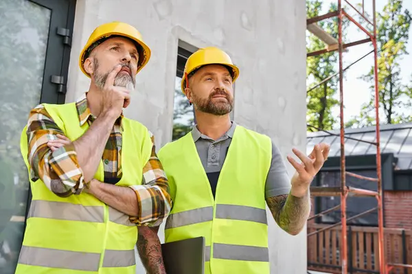 Dos trabajadores de la construcción de buen aspecto en chalecos de seguridad y cascos discutiendo sitio, constructores de cabañas - foto de stock
