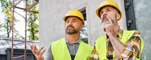 Dos trabajadores guapos en chalecos de seguridad y cascos discutiendo sitio, constructores de cabañas, pancarta - foto de stock