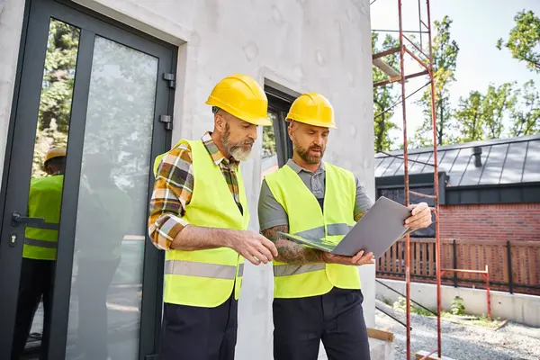 Stattliche bärtige Männer in Warnwesten und Helmen arbeiten mit Laptop auf der Baustelle, Bauarbeiter — Stockfoto