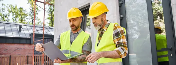 Handsome men in safety vests and helmets working with laptop on construction site, builders, banner — Stock Photo