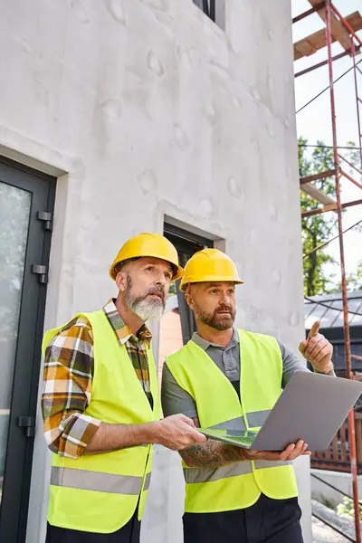 Handsome bearded men in safety vests and helmets working with laptop on construction site, builders — Stock Photo