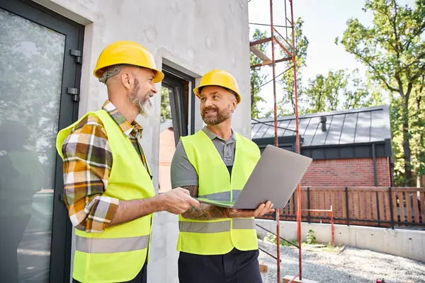 Cheerful bearded men in safety vests and helmets working with laptop on construction site, builders — Stock Photo