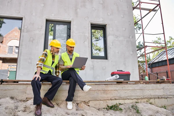 Appealing bearded cottage builders in safety helmets sitting on porch and working on laptop — Stock Photo