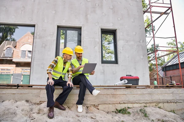 Attractive bearded cottage builders in safety helmets sitting on porch and working on laptop — Stock Photo