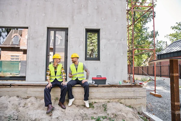 Joyful dedicated cottage builders in safety vests sitting on porch and looking at each other — Stock Photo