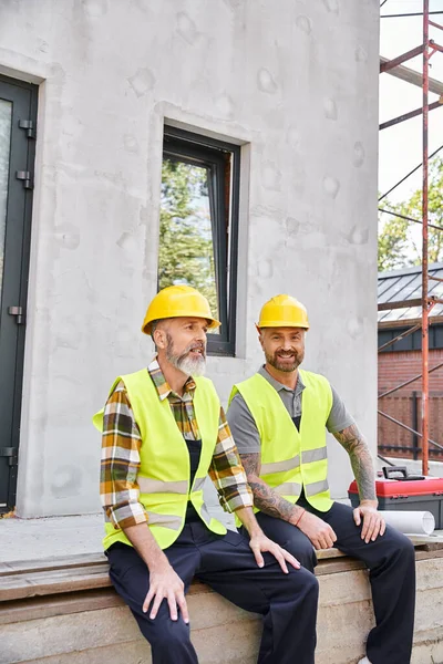 Cheerful hardworking cottage builders in safety helmets sitting on porch and looking at camera — Stock Photo