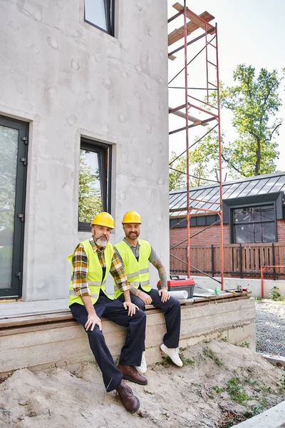 Веселі працьовиті будівельники котеджів у безпечних шоломах сидять на ганку і дивляться на камеру — стокове фото