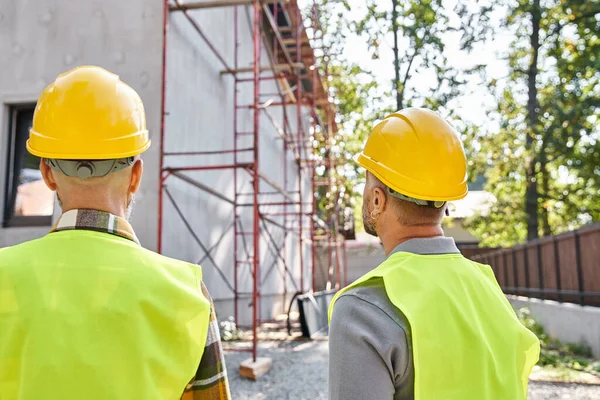 Back view of cottage builders in safety helmets looking at scaffolding on construction site — Stock Photo