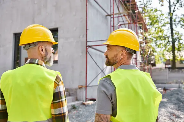 Handsome cottage builders in safety helmets posing next to scaffolding and looking at each other — Stock Photo