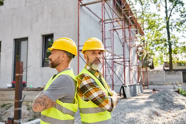 Handsome cottage builders in safety vests posing back to back near scaffolding and looking at camera — Stock Photo