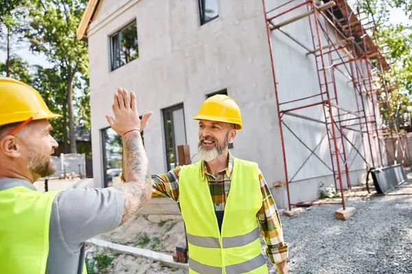 Two cheerful cottage builders in safety attires giving high five to each other near scaffolding — Stock Photo