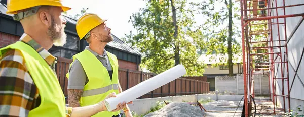 Two handsome cottage builders in safety helmets with blueprints working near scaffolding, banner — Stock Photo