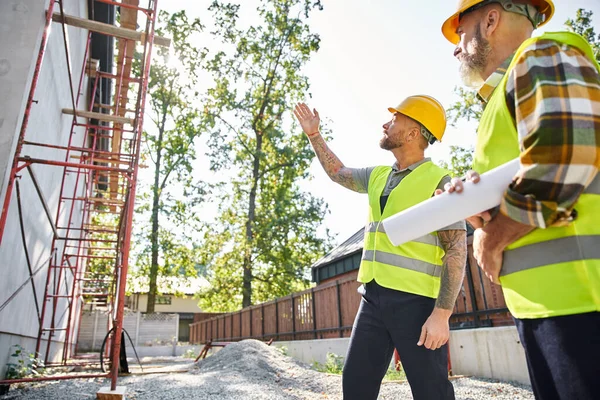 Two good looking cottage builders in safety helmets with blueprints working near scaffolding — Stock Photo