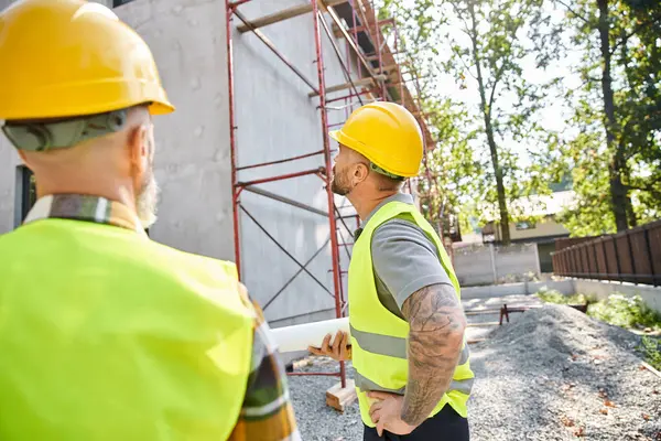 Focus on devoted cottage builder with blueprint posing next to his blurred colleague and scaffolding — Stock Photo