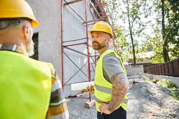 Focus on devoted cottage builder with blueprint posing next to his blurred colleague and scaffolding — Stock Photo