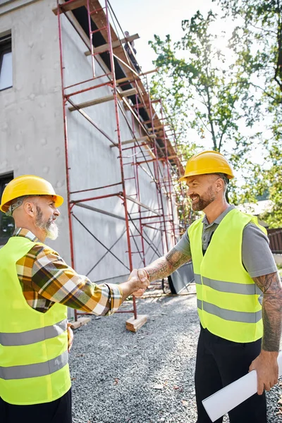 Trabajadores de la construcción alegres sacudiendo sus manos y sonriendo el uno al otro, constructores de cabañas - foto de stock