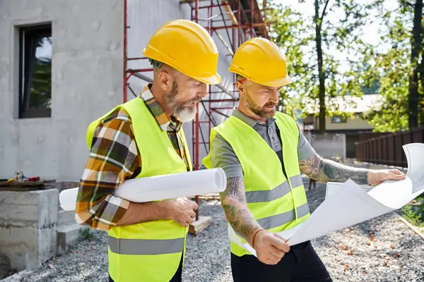 Appealing bearded cottage builders in safety vests working on their blueprints on construction site — Stock Photo
