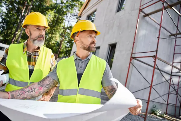 Appealing bearded cottage builders in safety vests working on their blueprints on construction site — Stock Photo
