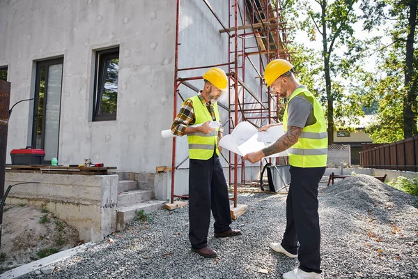 Attraktive bärtige Häuslebauer in Sicherheitswesten arbeiten auf der Baustelle an ihren Bauplänen — Stockfoto