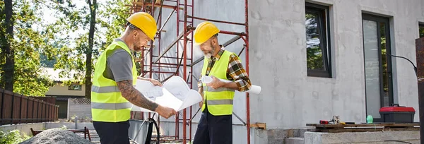 Attractive cottage builders in safety vests working on their blueprints on construction site, banner — Stock Photo