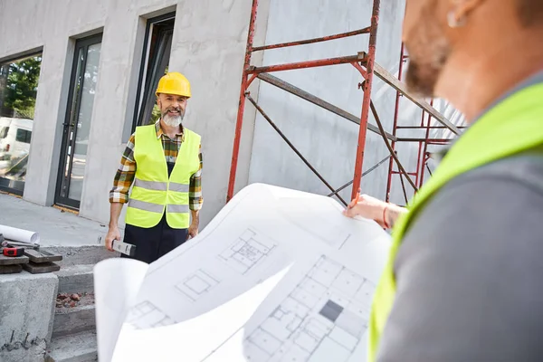 Focus on bearded jolly cottage builder with level looking at his blurred colleague with blueprint — Stock Photo