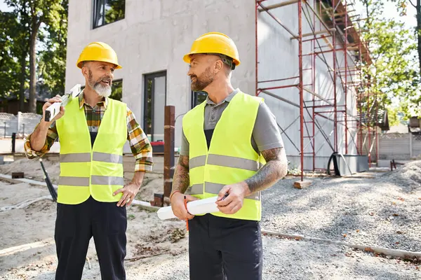Ansprechende freudige Bauarbeiter mit Ebene und Bauplan, die einander anschauen, Bauarbeiter — Stockfoto