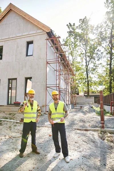Joyful cottage builders posing with blueprints and level near scaffolding and looking at camera — Stock Photo