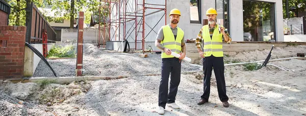 Beaux constructeurs de chalets avec des plans et un niveau près des échafaudages et en regardant la caméra, bannière — Photo de stock