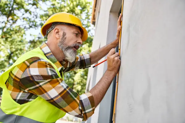 Handsome cottage builder in safety vest and helmet measuring window with tape while on site — Stock Photo