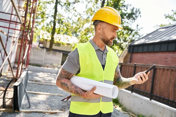 Handsome bearded construction worker with tattoos and blueprint looking at phone, cottage builder — Stock Photo