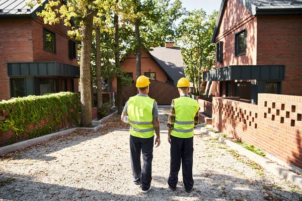 Back view of devoted construction workers in safety vests with blueprint on site, cottage builders — Stock Photo