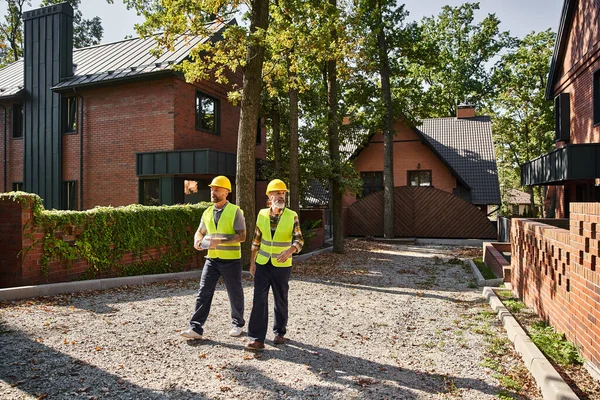 Good looking construction workers in safety helmets walking and discussing site, cottage builders — Stock Photo
