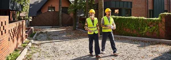 Beaux travailleurs de la construction en gilets de sécurité marchant et discutant du site, constructeurs de chalets, bannière — Photo de stock