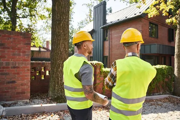 Back view of dedicated construction workers in safety vests with blueprint on site, cottage builders — Stock Photo