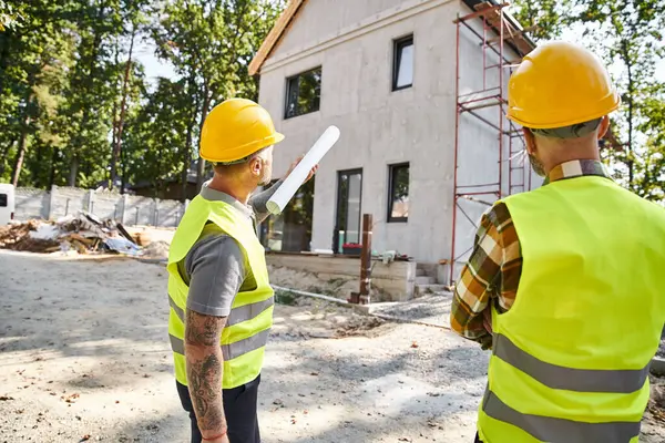 Back view of devoted cottage builders in safety helmets with blueprint discussing their work on site — Stock Photo