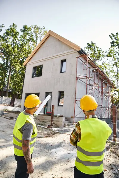 Back view of devoted cottage builders in safety helmets with blueprint discussing their work on site — Stock Photo