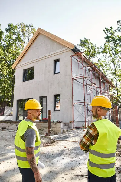 Good looking devoted cottage builders in safety helmets with blueprint discussing construction site — Stock Photo