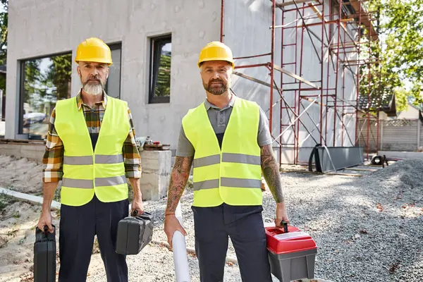 Due uomini attraenti in caschi di sicurezza e giubbotti in posa con cassette degli attrezzi e cianografia, costruttori di case — Foto stock