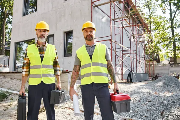 Dos hombres atractivos en cascos de seguridad y chalecos posando con cajas de herramientas y planos, constructores de cabañas - foto de stock